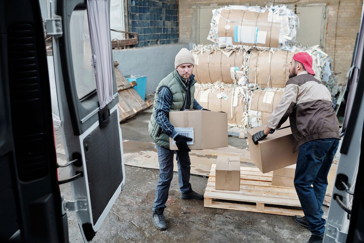 Twee mannen laden dozen van een pallet in een wagen, gezien vanuit de laadruimte