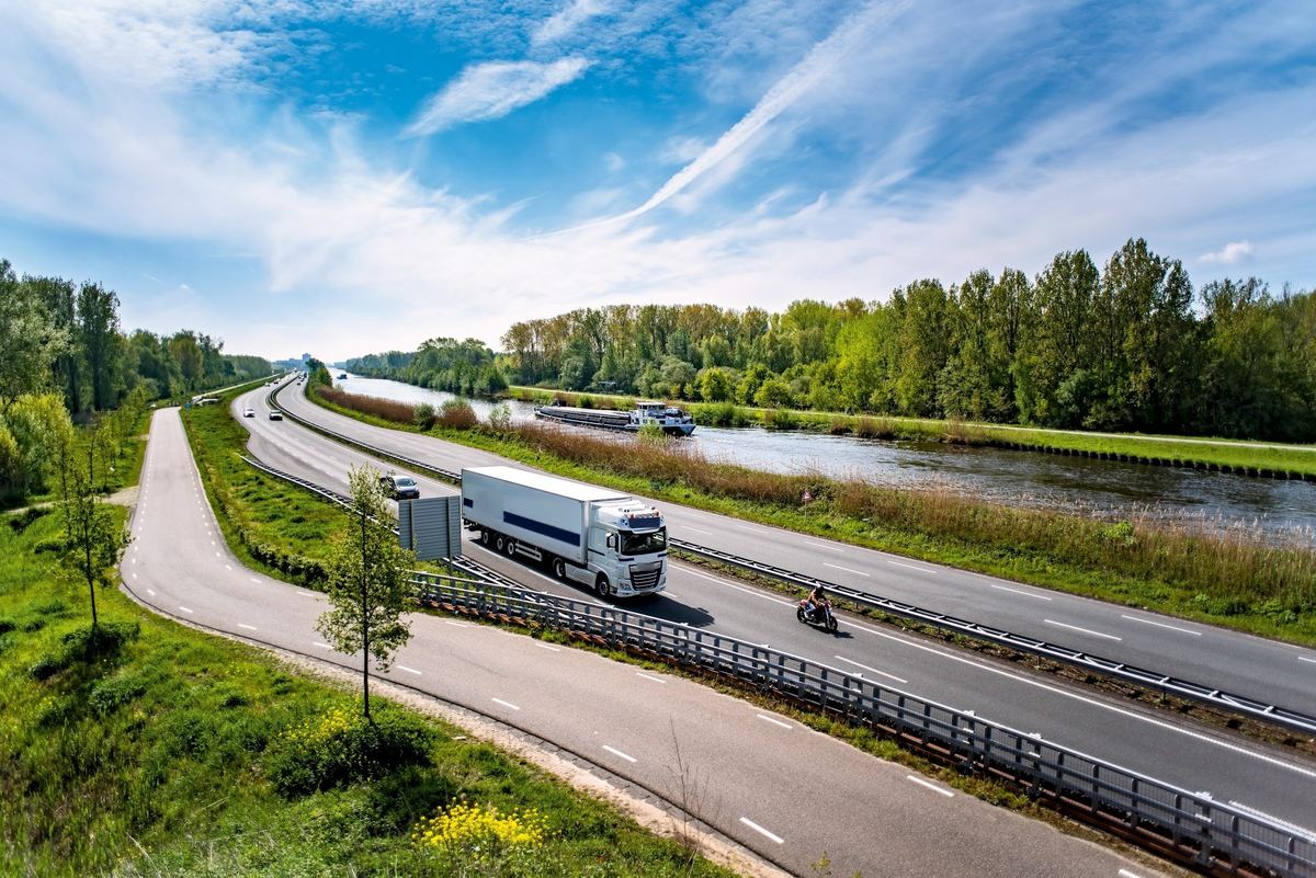 Een witte vrachtwagen met oplegger rijdt over een snelweg langs een kanaal, met bomen en een binnenschip op het water